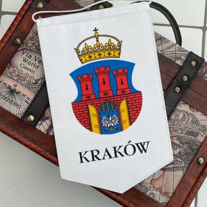 May include: White pennant flag featuring the coat of arms of Kraków, Poland. The emblem includes a blue shield with three red towers, a golden crown, and the city's name, "KRAKÓW," in black. The flag is displayed on a decorative wooden chest.