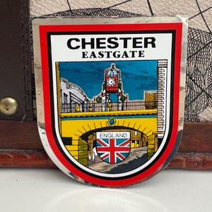 Op de afbeelding: Een rechthoekige sticker met een rode rand toont de tekst "CHESTER EASTGATE" boven een kleurrijke illustratie van een klokkentoren en brug. De Union Jack-vlag is zichtbaar, met het woord "ENGLAND" eronder.