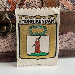 May include: A white fabric patch with a gold and black coat of arms featuring a green wreath, a red figure holding a stick, and the text "MONNIKENDAM".