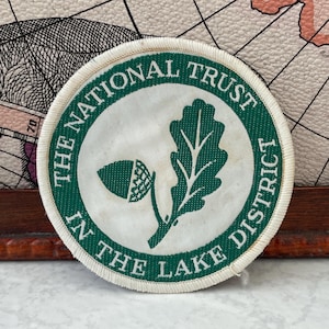 May include: A circular embroidered patch featuring a white centre with a green oak leaf and acorn graphic. The green border contains the white text THE NATIONAL TRUST IN THE LAKE DISTRICT. The patch has a white stitched edge and a flat design.