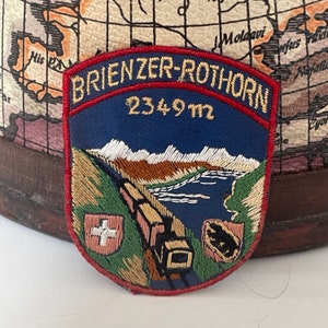 May include: A blue and red embroidered patch with the text "BRIENZER-ROTHORN 2349 m" and a depiction of a train traveling through a mountain landscape. The patch features a Swiss flag and a small brown bear.