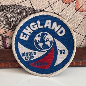 May include: A circular embroidered patch featuring a blue background with white text that reads ENGLAND WORLD CUP '82 and SPAIN. The center displays a white globe graphic, and the patch is finished with a white stitched border edge.