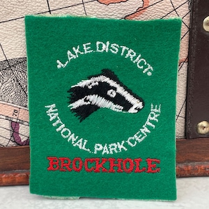 May include: A rectangular green felt patch featuring white embroidered text that reads Lake District National Park Centre, a black and white badger head illustration, and the word BROCKHOLE embroidered in red capital letters at the bottom.