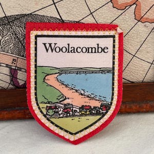 以下が含まれることがあります： 上部に「Woolacombe」の文字が入った赤と白の刺繍パッチ。青い海、砂浜、小さな家々が描かれたビーチの風景が特徴です。黄色いステッチの縁取りがあります。
