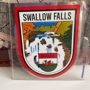 Op de afbeelding: Een rode, witte en groene vintage sticker met de tekst "SWALLOW FALLS WALES" en een rood drakenwapen. De sticker toont een cartoonillustratie van een waterval in een bos.