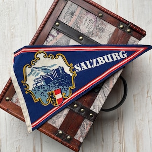 May include: A blue pennant with the word "SALZBURG" in white, featuring an illustration of a castle and mountains. The pennant has red and white stripes and is displayed on a vintage wooden trunk.