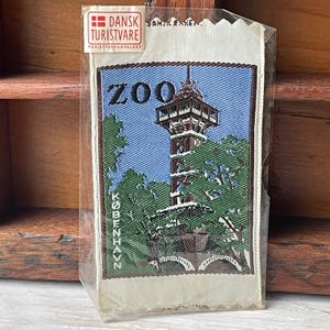 May include: A vintage souvenir patch featuring a tall tower with the word "ZOO" and "KOBENHAVN" in green and white text. The patch is in a clear plastic sleeve with the "DANSK TURISTVARE" logo.