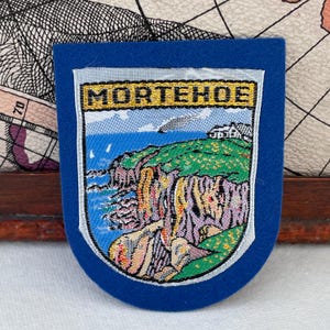 Puede incluir: Un parche de fieltro azul con una imagen bordada de un paisaje costero. La imagen presenta acantilados, el océano y un edificio, con la palabra "MORTEHOE" en la parte superior. El parche tiene forma de escudo.