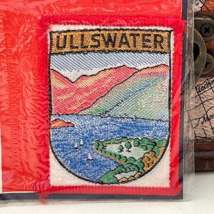 May include: A red, white and blue embroidered patch with the text "ULLSWATER" in gold lettering. The patch features a scenic landscape with mountains, a lake and trees.