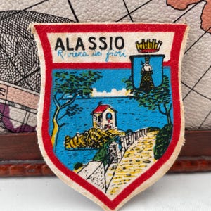 May include: A vintage fabric patch with a shield design featuring the text "ALASSIO Riviera dei fiori". The patch has a red border and depicts a coastal scene with a building, trees, and a pathway. The background is a map.