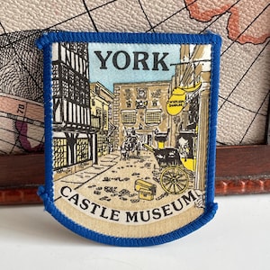 May include: A rectangular patch with a blue border depicting a street scene in York, England. The patch features the text "YORK" at the top and "CASTLE MUSEUM" at the bottom. The illustration shows buildings, a horse-drawn carriage, and cobblestone streets.