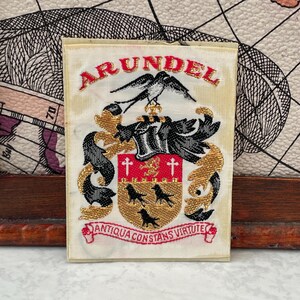 May include: A rectangular fabric patch featuring the Arundel coat of arms embroidered in gold, black, and red thread. The design includes a bird crest, a shield with three black birds, and a banner with the Latin text ANTIQUA CONSTANS VIRTUTE.