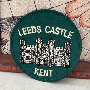 Può includere: Una toppa rotonda verde con testo ricamato bianco e un'illustrazione di un castello. Il testo recita "LEEDS CASTLE" sopra l'immagine del castello e "KENT" sotto. Il castello è bianco con dettagli blu e marroni.