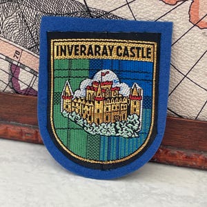 Op de afbeelding: Een blauwe patch met bovenaan de tekst "INVERARAY CASTLE". De patch toont een gedetailleerde afbeelding van een kasteel met rode daken en gouden accenten, tegen een groen-blauwe geruite achtergrond. De patch is goudkleurig omrand.