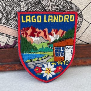 May include: A shield-shaped embroidered patch featuring the text LAGO LANDRO in yellow. The design depicts a blue lake, green trees, red mountains, a brown cabin with the text RAST, and a white edelweiss flower with red accents at 1410 m.