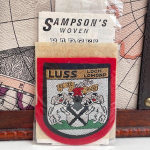 May include: A woven badge from Sampson's, featuring the text "LUSS LOCH LOMOND" and a coat of arms with two dogs, a rose, and a shield. The badge has a red border and is packaged with a paper backing.