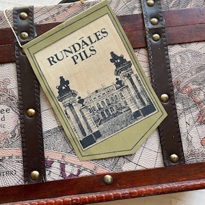 May include: A vintage-style travel trunk with a map design and dark brown leather straps. A green tag with the text "RUNDĀLES PILS" and a black illustration of a building is attached to the trunk.