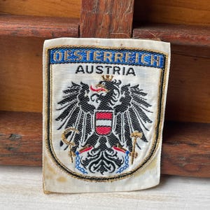 May include: Embroidered patch featuring the Austrian coat of arms. The patch has a white background with the words "OESTERREICH" and "AUSTRIA" in blue. The coat of arms includes a black eagle, a red and white shield, and gold accents.