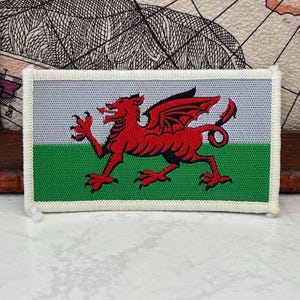 以下が含まれることがあります： ウェールズの旗をあしらった長方形の刺繍ワッペン。上半分が白、下半分が緑の水平な二色背景で、中央には大きく詳細な赤いドラゴンの刺繍が施されています。縁は白い糸で丁寧に縫い合わされており、全体的にしっかりとした作りになっています。