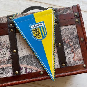 May include: A blue and yellow triangular pennant with a white crest featuring a lion and the word "LEIPZIG" in black lettering. The pennant is hanging from a wooden chest with a map design.