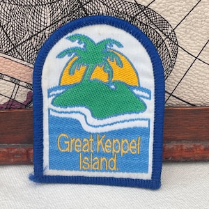Op de afbeelding: Een rechthoekige patch met een blauwe rand toont een tropisch eiland. Een groen eiland met een palmboom tegen een gele zon en blauw water. De tekst "Great Keppel Island" is geel.