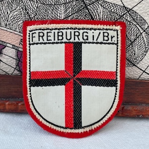 Puede incluir: Parche en forma de escudo con borde rojo y centro blanco. El centro presenta un diseño de cruz en rojo y negro. El texto "FREIBURG i/Br." está impreso en la parte superior en negro.