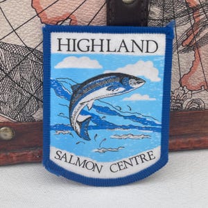 Può includere: Una toppa rettangolare con bordo blu presenta le parole "HIGHLAND SALMON CENTRE". Un salmone che salta è raffigurato sopra le parole, su uno sfondo di cielo blu con nuvole bianche. La toppa è bianca con dettagli blu e neri.