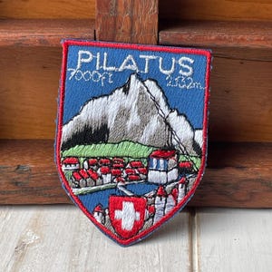 May include: Embroidered patch with a blue background featuring the word "PILATUS" and a mountain scene. The patch includes the text "7000ft" and "2133m". A Swiss cross is at the bottom.