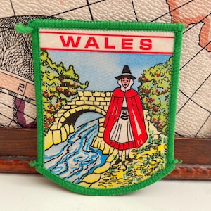 May include: A green-edged pennant displaying "WALES" in red. The image shows a figure in a red cloak and hat beside a stone bridge over a river. The background includes trees and a blue sky.