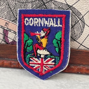 Könnte beinhalten: Blaues, schildförmiges Abzeichen mit dem weißen Wort "CORNWALL". Es zeigt eine Figur, die ein Horn spielt, mit der Flagge des Union Jack am unteren Rand. Das Abzeichen ist mit roten Nähten und einem weißen Rand versehen.