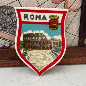 May include: A shield-shaped patch featuring the text ROMA in black letters. The design includes a depiction of the Colosseum in brown and tan tones, set against a light blue sky. A small red crest with a golden wolf emblem is positioned at the top right.