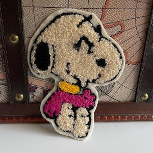 Könnte beinhalten: Gestickter Aufnäher mit einem Cartoon-Hund mit cremefarbenem Körper, schwarzen Ohren und Augen sowie einem rosa Schal. Der Hund trägt ein gelbes Halsband. Der Aufnäher befindet sich auf einem cremefarbenen Hintergrund.