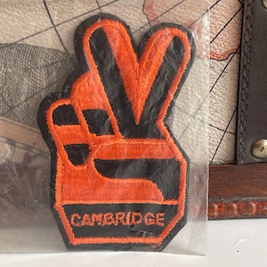 Op de afbeelding: Een geborduurde patch met een hand die een vredesteken maakt in feloranje draad tegen een zwarte achtergrond. Het woord CAMBRIDGE is in oranje hoofdletters geborduurd aan de onderkant van de patch, die in doorzichtig plastic is verpakt.