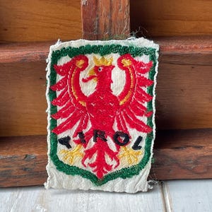 Könnte beinhalten: Gestickter Aufnäher mit einem roten Adler mit goldener Krone und gelben Krallen. Der Adler befindet sich auf weißem Grund, umrahmt von einem grünen Rand. Der Text "TIROL" steht darunter.