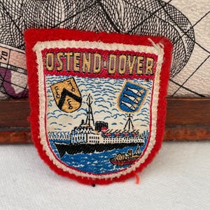 May include: A vintage embroidered patch in red, white, and blue, featuring the words "OSTEND-DOVER". The patch depicts a ship, waves, and two crests. It has a rectangular shape with rounded corners.