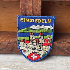 Puede incluir: Parche bordado con forma de escudo azul, con la palabra "Einsiedeln" en dorado. El parche muestra una ilustración detallada de una iglesia con tejados rojos, montañas y un emblema de la bandera suiza.