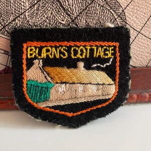 May include: Embroidered patch with the text "BURN'S COTTAGE" in gold lettering. The patch features a detailed image of a cottage with a tan roof, green accents, and a black border with orange trim. The patch is on a black background.