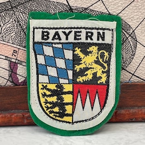 Könnte beinhalten: Ein rechteckiger Stoffaufnäher mit grünem Rand und der Aufschrift BAYERN am oberen Rand. Das Wappen ist in vier Felder unterteilt: blau-weiße Rauten, ein gelber Löwe auf Schwarz, schwarze Löwen auf Gelb und Rot mit weißen Spitzen.