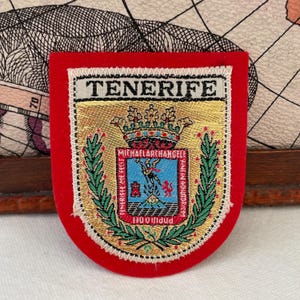 Puede incluir: Parche rojo con la palabra "TENERIFE" en la parte superior. El parche presenta un emblema detallado con una corona, un escudo y las palabras "MICHAELARCHANGELE" y "POPULI DEI". El emblema está rodeado de hojas verdes.