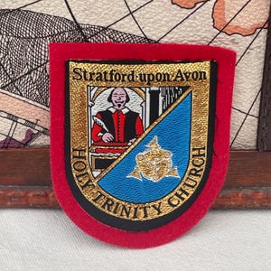 May include: A red, shield-shaped patch with gold embroidered text and details. The patch features the text "Stratford-upon-Avon" and "Holy Trinity Church". A portrait of a man in a red and black outfit is in the center.