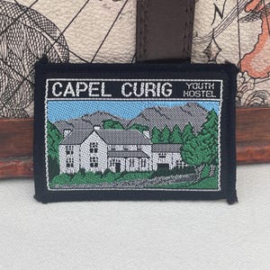 May include: A rectangular patch with the text "CAPEL CURIG YOUTH HOSTEL" in white. The patch features a detailed illustration of a white building with green trees and mountains under a blue sky. The patch has a black border.