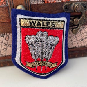 May include: A blue and white felt patch with the word "WALES" in gold lettering. The patch features a red shield with three white feathers and the text "Ich Dien" in gold lettering.