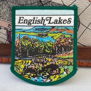 Op de afbeelding: Een rechthoekige geborduurde patch met een donkergroene rand en de tekst English Lakes in het zwart. Het ontwerp toont een kleurrijk landschap met een stenen brug op de voorgrond, groene bomen, een blauw meer en heuvels.