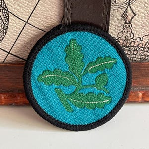 May include: A circular embroidered patch featuring a bright turquoise background with a dark green oak leaf design in the centre. The patch has a thick black stitched border and is attached to a dark brown leather-like strap at the top.