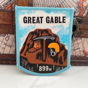 May include: A rectangular patch with a light blue border features the text "GREAT GABLE" above a brown mountain illustration. The illustration includes a pickaxe, hiking boots, and an orange helmet. The patch also displays "899M".