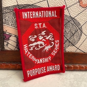 Op de afbeelding: Een rechthoekige rode patch met witte tekst en een centraal embleem. De tekst luidt "INTERNATIONAL S.T.A. WATERMANSHIP SERIES PORPOISE AWARD". Het embleem toont een witte bruinvis. De patch heeft een rode rand.