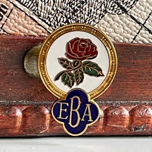 Puede incluir: Un pin de oro con un círculo blanco que contiene una rosa roja y hojas verdes. El círculo está rodeado por un borde dorado. Debajo del círculo hay un trébol azul con las letras "EBA" en oro.