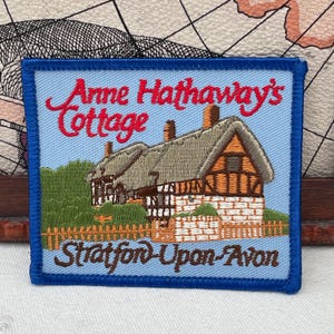 May include: Embroidered patch featuring Anne Hathaway's Cottage in Stratford-Upon-Avon. The patch has a blue border and a light blue background. The cottage is depicted with a thatched roof, brick chimney, and a wooden fence.