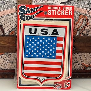 May include: A red, white and blue American flag sticker with the words "U.S.A." in black lettering. The sticker is in a shield shape and has a white border. The sticker is packaged in a clear plastic sleeve with a white backing card that says "Sampson Souvenir Double Sided Sticker".