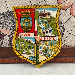 Könnte beinhalten: Ein schildförmiger Stoffaufnäher mit gelbem Kordelrand. Die linke Seite zeigt ein rotes Wappen mit goldener Krone und Adler. Die rechte Seite stellt Tivoli und die Villa d'Este mit Brunnen dar. Der Text lautet TIVOLI und VILLA D'ESTE.